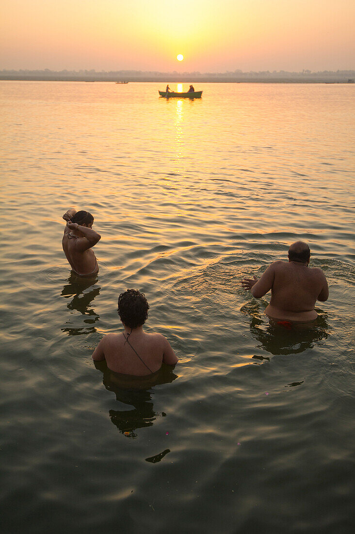 Three devotees wash in the Ganges river waters while a boat passes and the sun is rising in the background