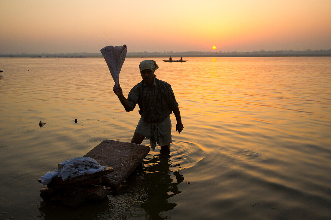 Silhouette of a man washing cloths in the Ganges river at sunrise white boats pass and the sun is rising in the background, Varanasi, India