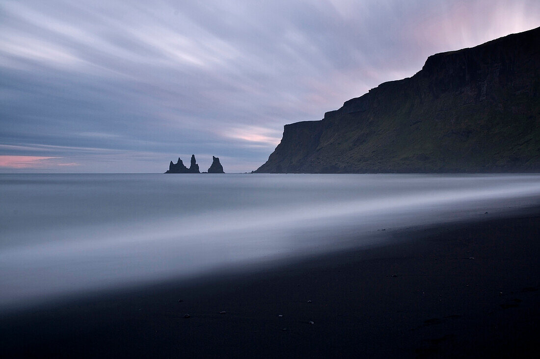 Long exposure of the ocean on a cloudy dusk on the black sand beach in front of the rock formation of Vik, Iceland
