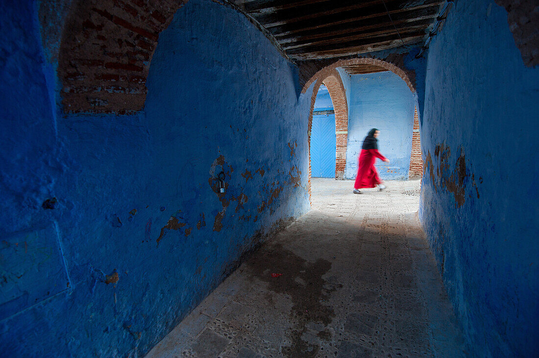 A woman with red dress walks through the blue colored streets of the blue city of Chefchaouen, Morocco