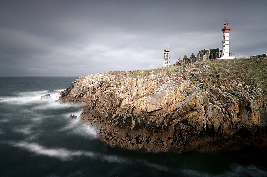 The ocean crushes onto the cliffs of the the lighthouse of Pointe Saint Mathieu in the dim light of a stormy morning in Brittany, France