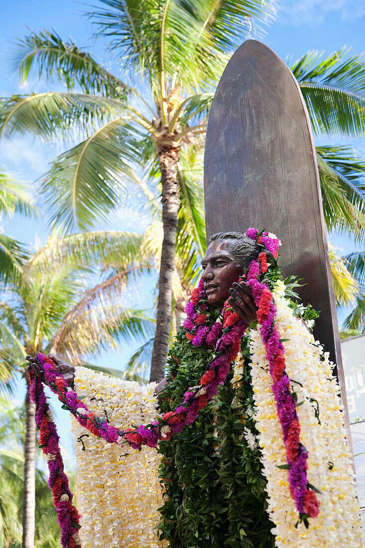 'Statue of a man covered in floral leis standing against a surfboard; Honolulu, Oahu, Hawaii, United States of America'