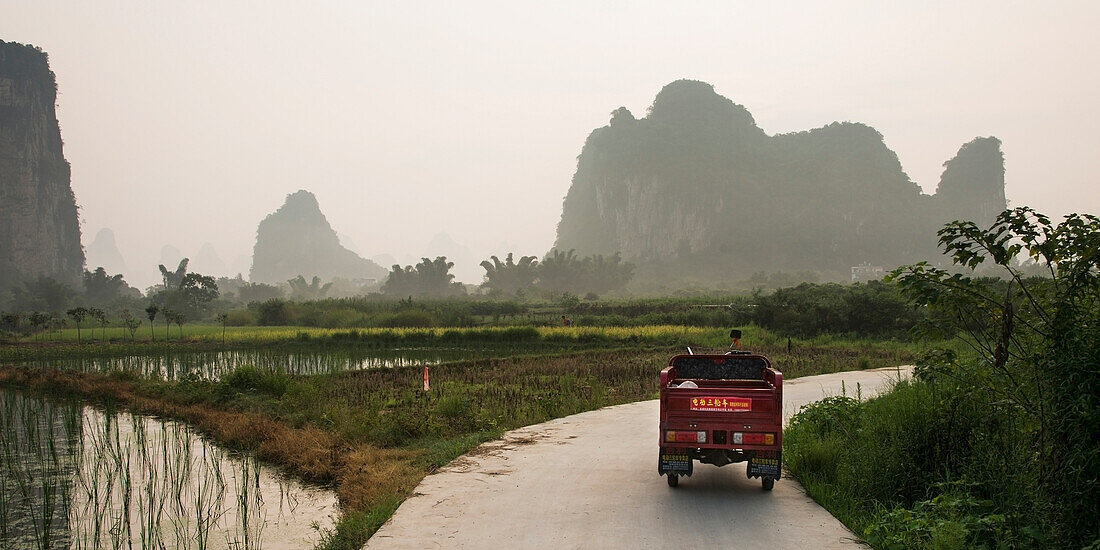 'A Truck Travels Down A Road At Sunset Beside Peaked Mountains And Water; Yangshuo, Guangxi Province, China'