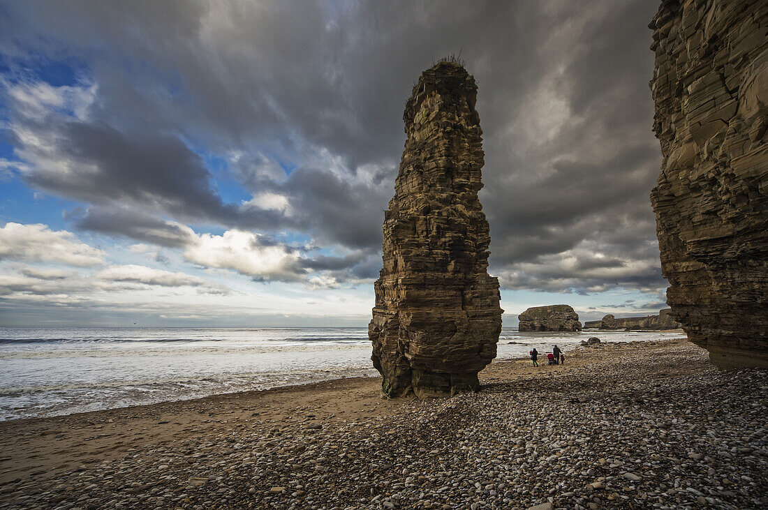 'A family walks on the beach along the water with a tall rock column; South Shields, Tyne and Wear, England'