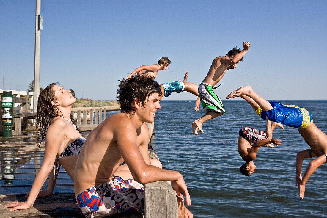 Young Couple Sitting on Edge of Pier With Group of Boys Jumping in Water