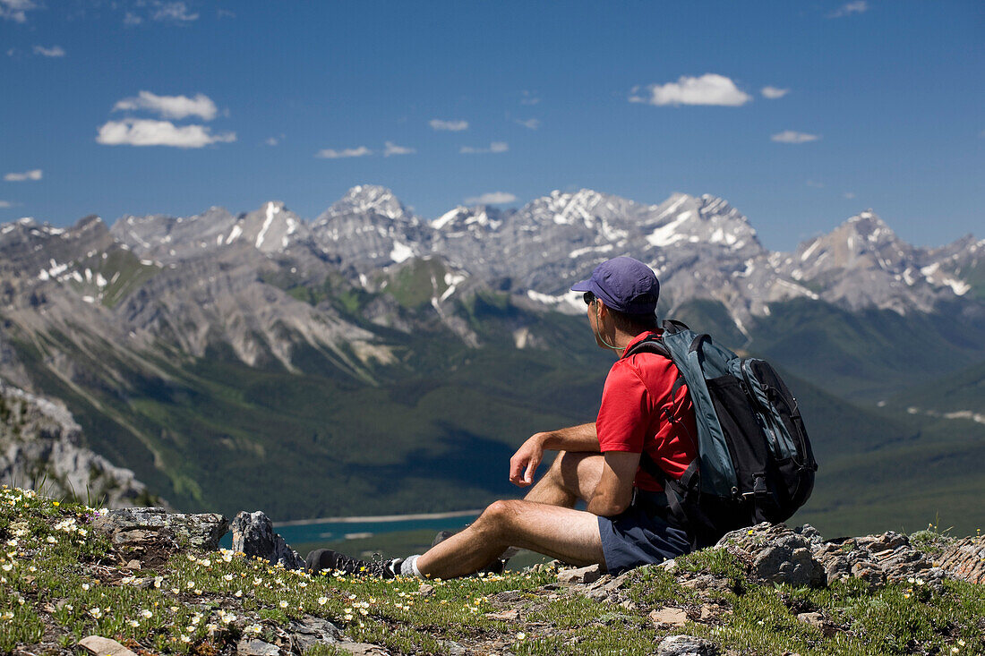 'Male Hiker Sitting On Top Of A Ridge Overlooking A Mountain Valley And Lake; Alberta, Canada'