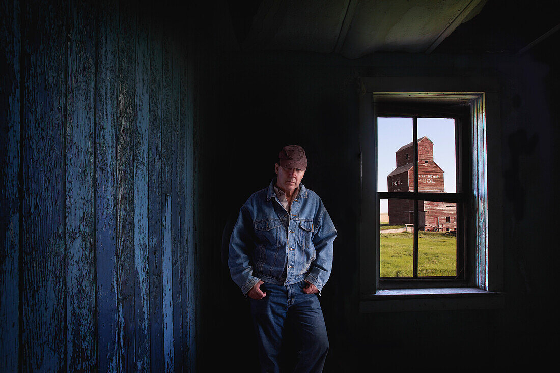 'Portrait Of A Farmer/Rancher In An Old Abandoned Ghost Town Store With A Grain Elevator Seen Out The Window; Bents, Saskatchewan, Canada'