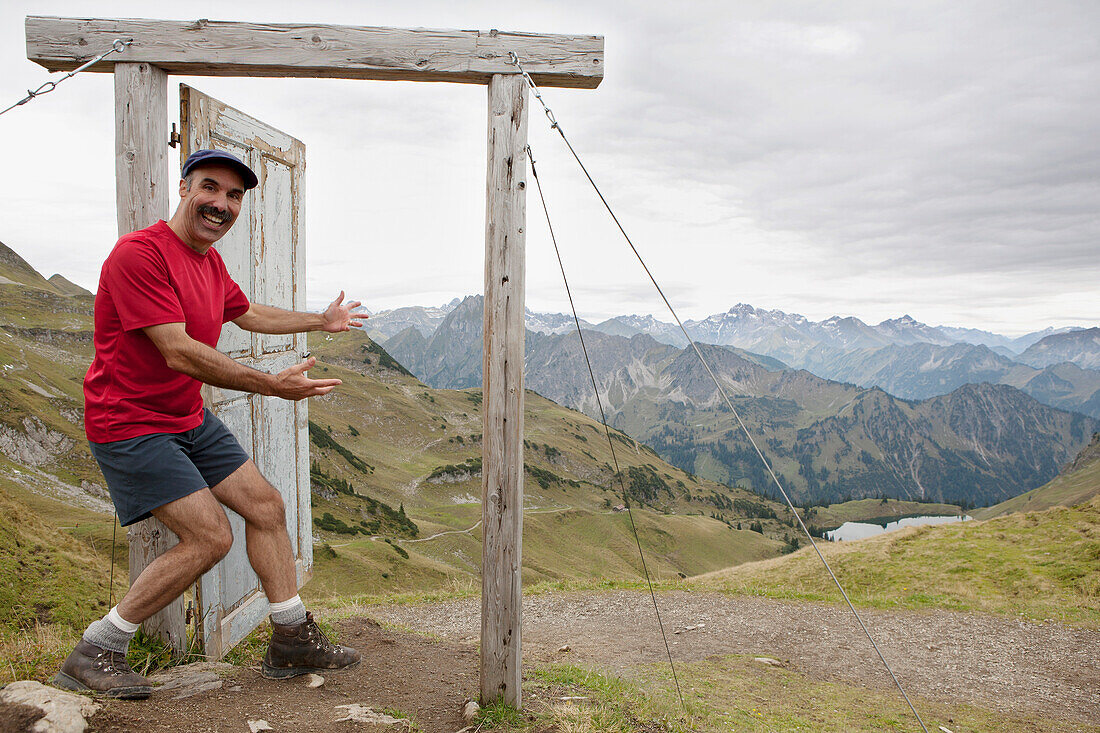 'Male Hiker On Top Of A Mountain Stepping Into An Open Wooden Door And Frame Overlooking A Mountain Valley, Lake And Mountain Range; Oberstdorf, Germany'