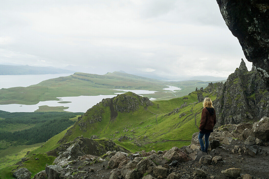 'A woman stands along on a rocky ridge looking out over the landscape of hills and water; Skye, Scotland'