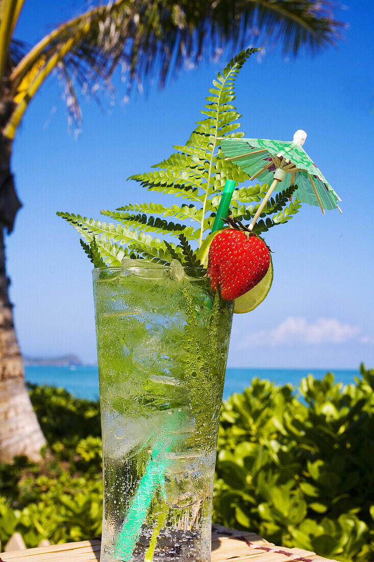 A Tropical Cocktail Garnished With Fruit And Green Fern Leaves In An Outdoor Setting.