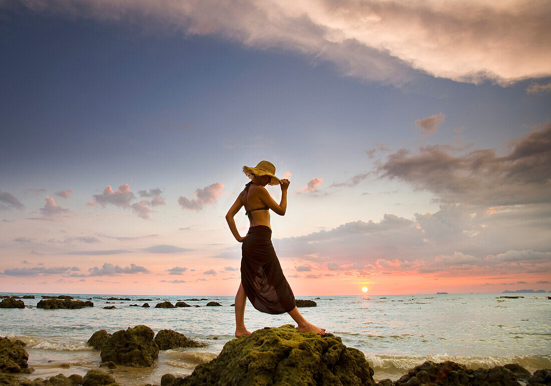 'A Woman Wearing A Hat And Sarong Stands On The Beach Of A Tropical Island At Sunset; Koh Lanta, Krabi Province, Thailand'
