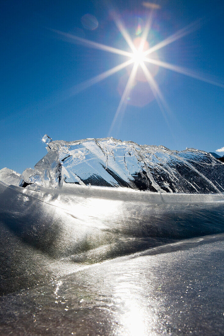 'Ice Formations On A Frozen Lake Shoreline In The Mountains With A Sunburst And Blue Sky; Alberta, Canada'