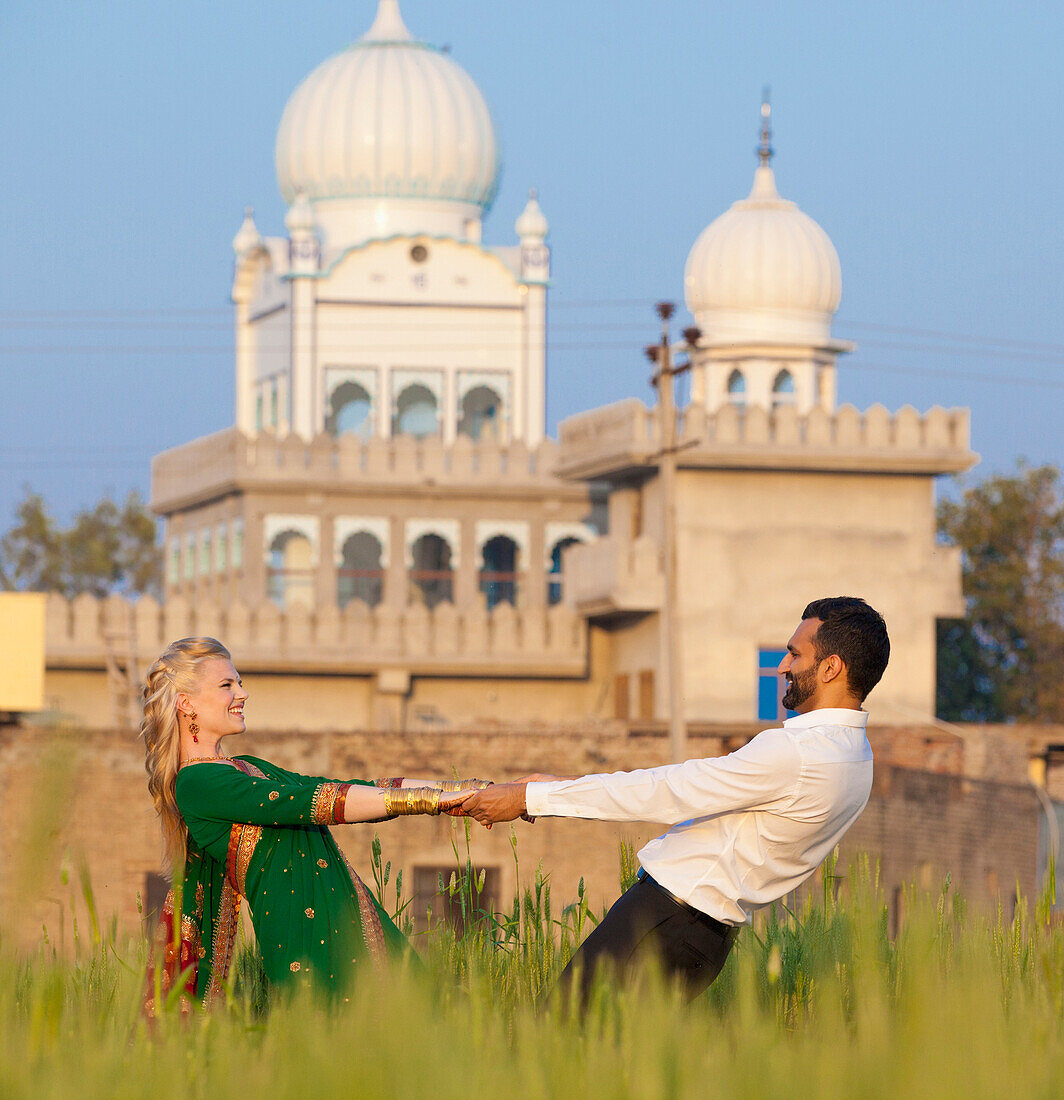 'A Mixed Race Couple Holding Hands In A Field With A Temple In The Background; Ludhiana, Punjab, India'
