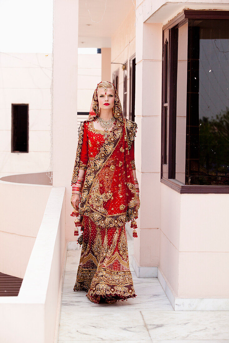 'A Bride In An Ornate Red And Gold Gown Walking Down A Corridor; Ludhiana, Punjab, India'