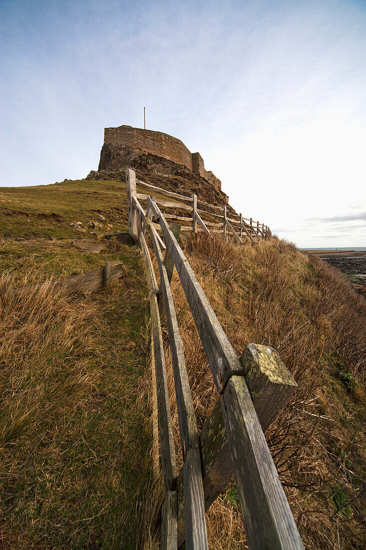 'A Weathered Wooden Fence Along The Water's Edge With A Building In The Background; Lindisfarne, Northumberland, England'