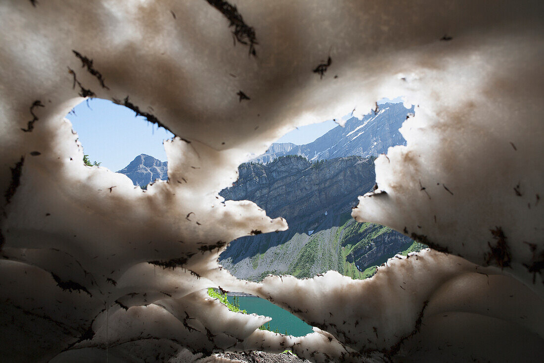 'Underneath a melting snow pack with gaps showing mountains and lake with blue sky in kananaskis provincial park;Alberta canada'