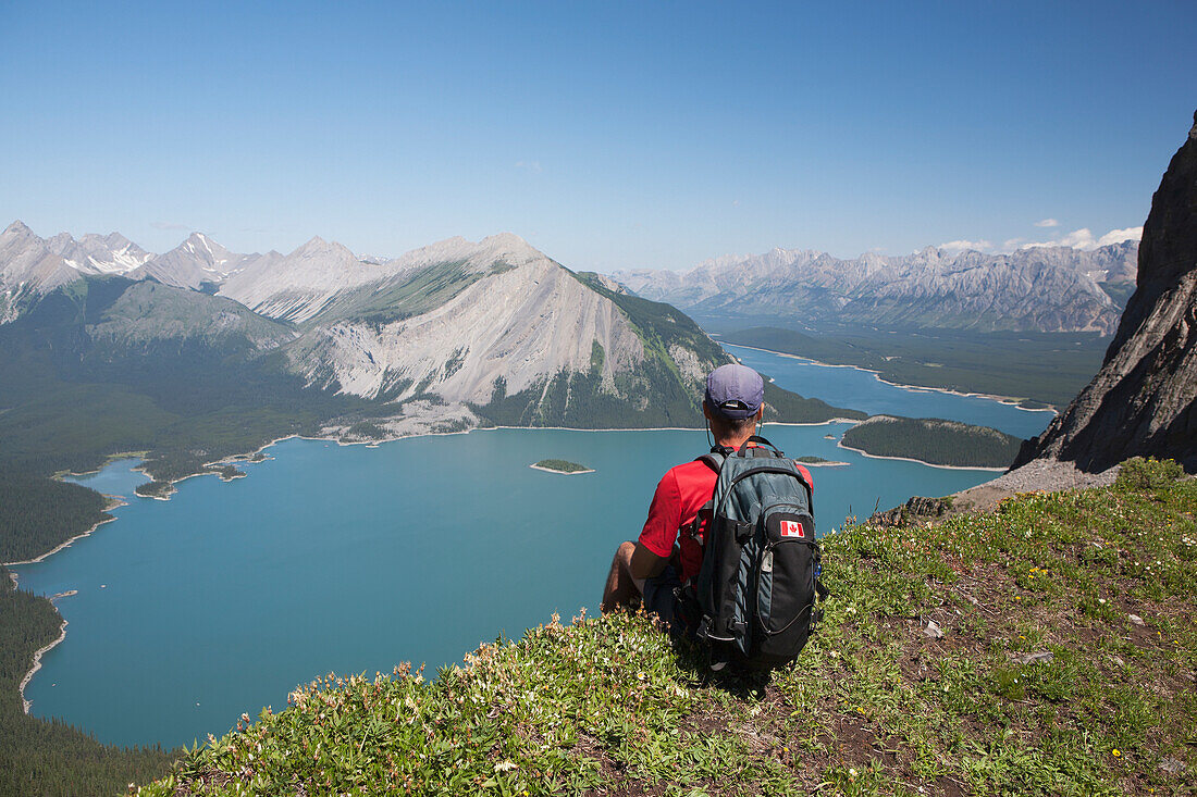 'Male hiker sitting on a mountain ridge overlooking an emerald lake and mountains below with blue sky in kananaskis provincial park;Alberta canada'