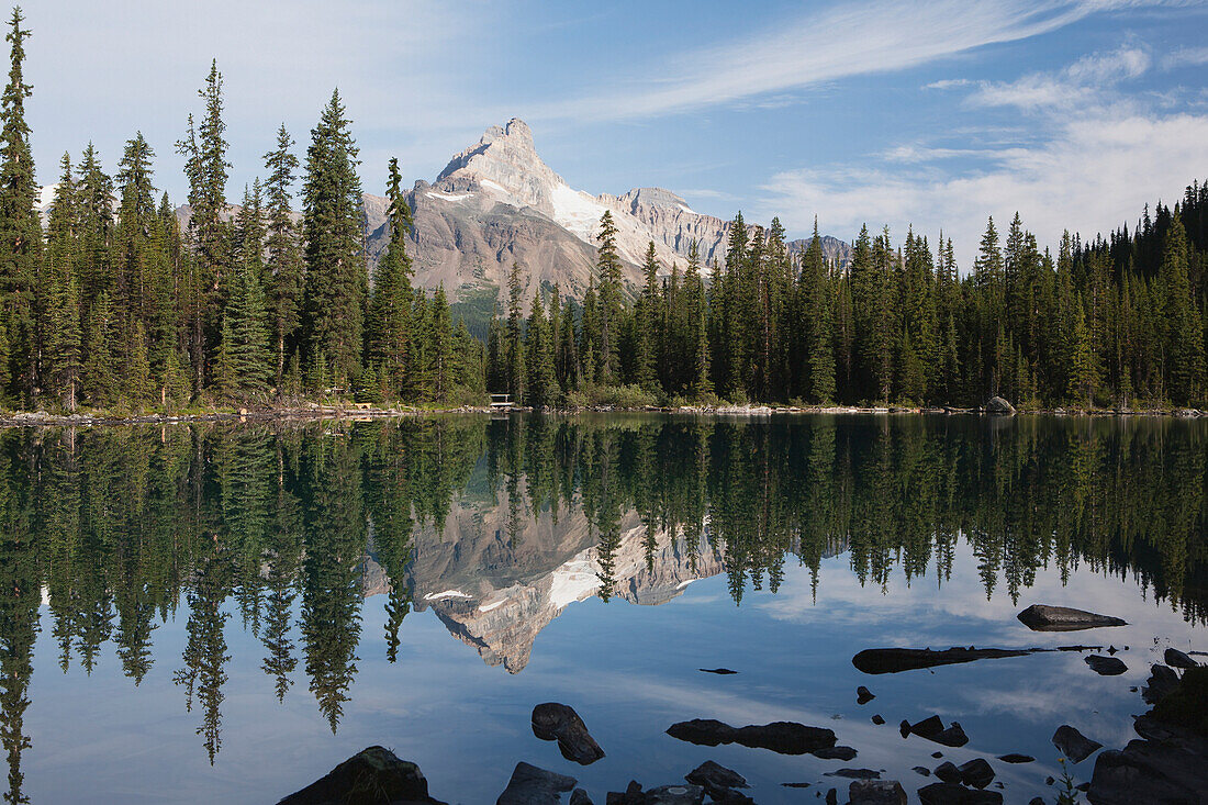 'Mountain lake reflecting mountain peak at sunrise with trees framing lake and blue sky with clouds;Field british columbia canada'