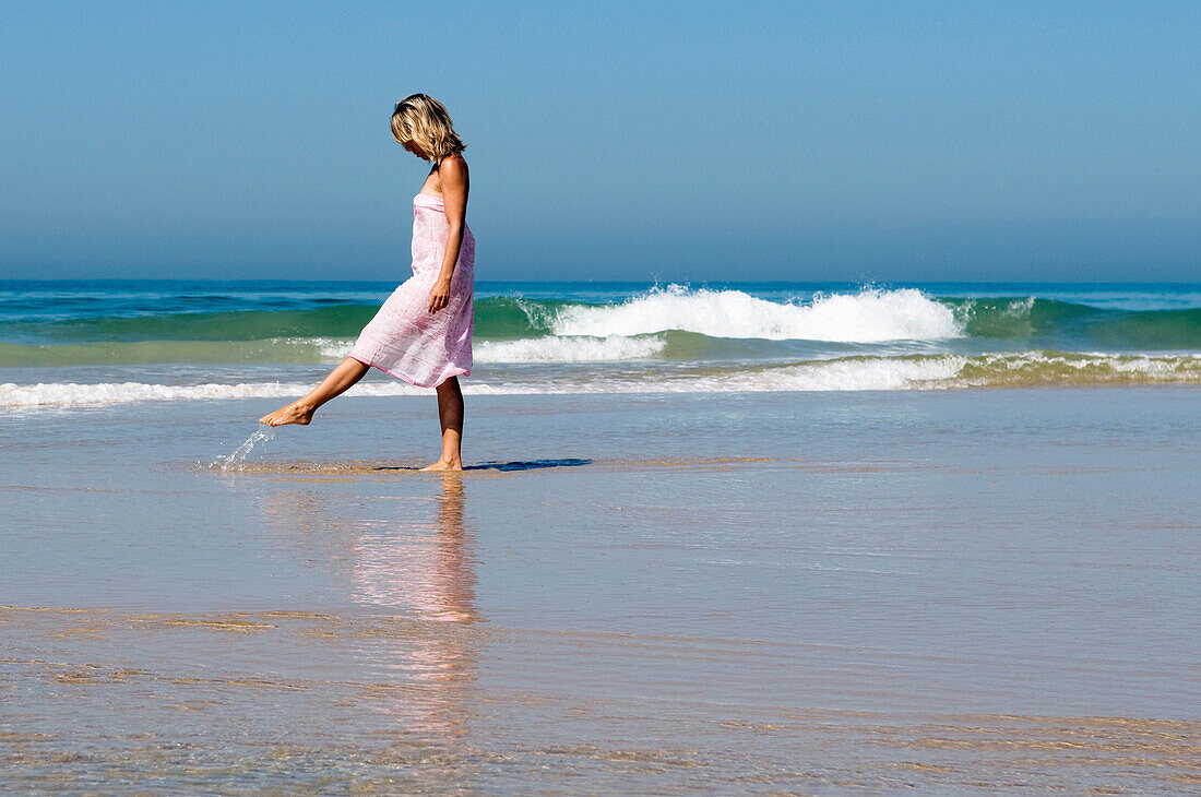 Young Woman Walking Along The Beach Kicking Up Water