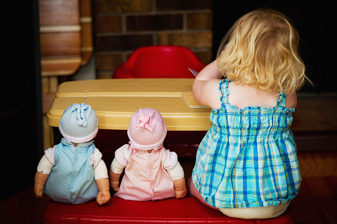 'A toddler sits at a play table with her two dolls; Alberta, Canada'