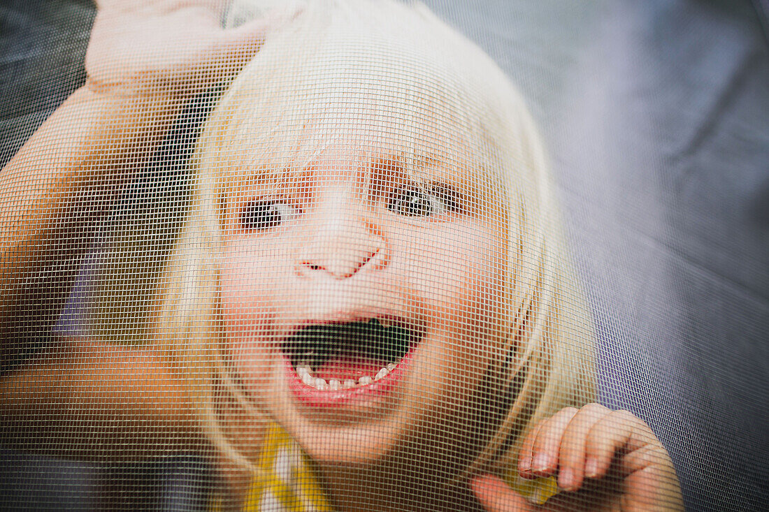 'A young girl with a silly expression on her face viewed through a window screen; Peachland, British Columbia, Canada'