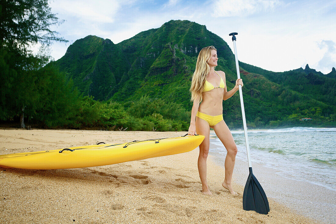 'A young woman in a yellow bikini stands at the water's edge with a paddleboard and paddle; Kauai, Hawaii, United States of America'