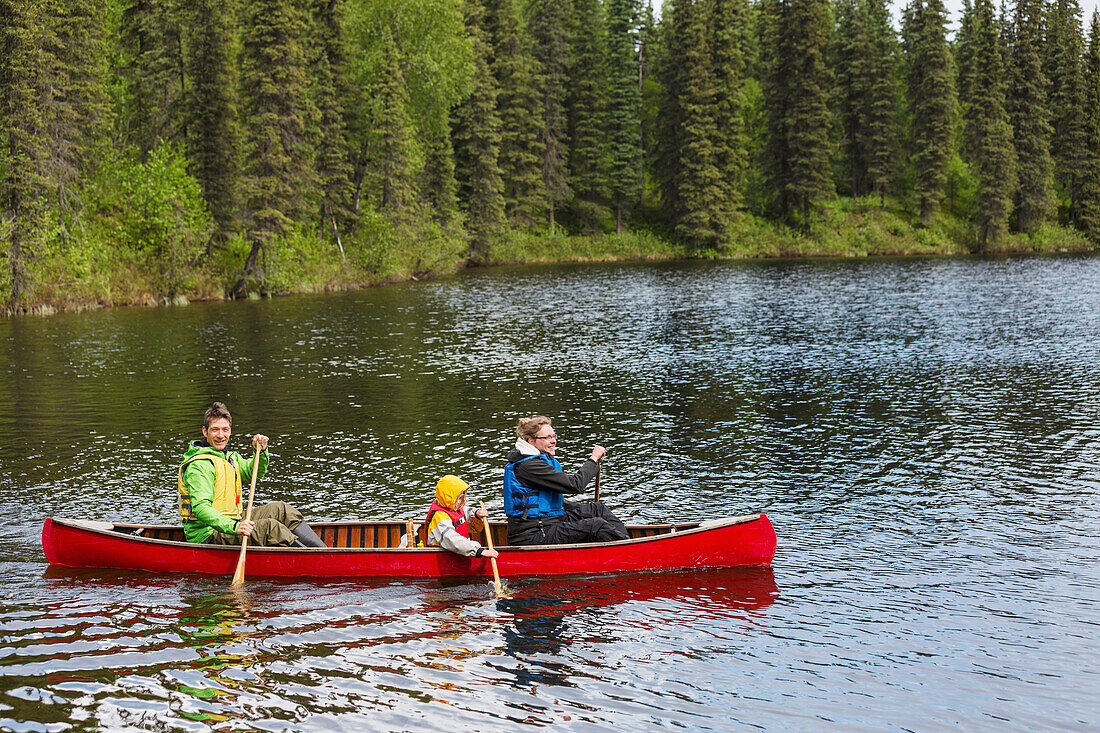 'Family in red canoe on Byers Lake with green tree covered shoreline, Denali State Park; Alaska, Alaska, United States of America'