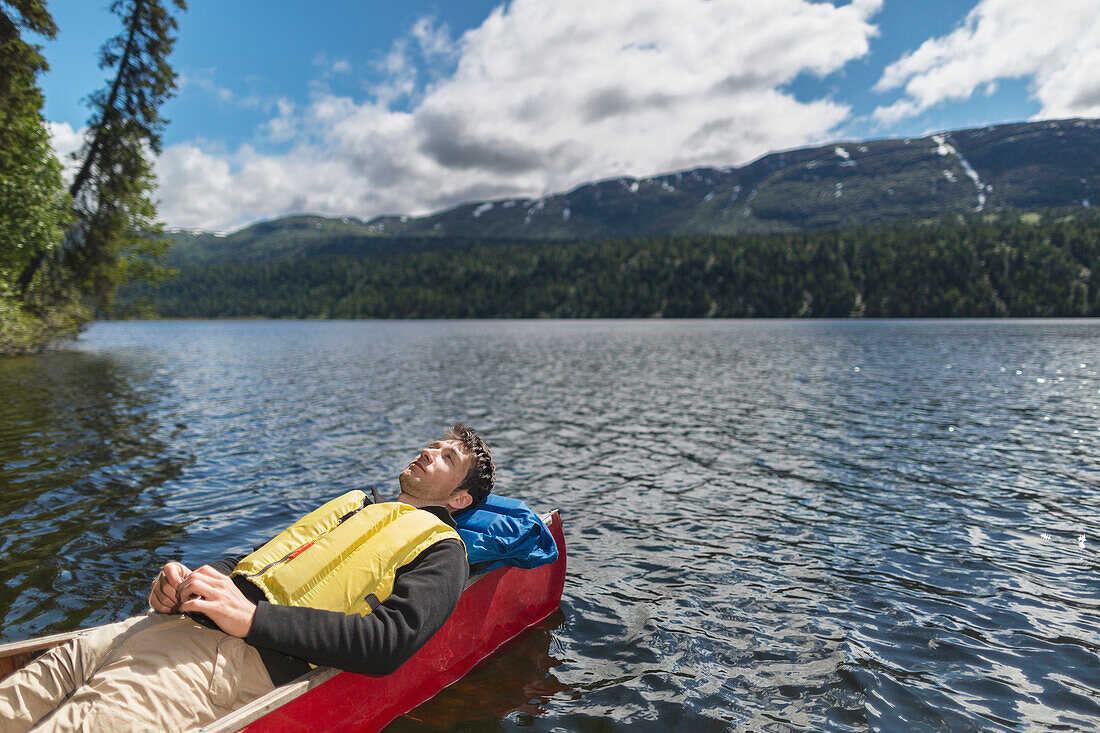 'Man reclined in canoe and resting in the sunshine on Byers Lake with forested foothills in the background, Byers lake campground, Denali State Park; Alaska, United States of America'