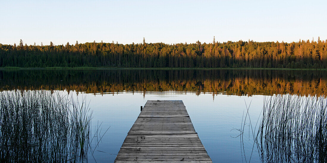 'A wooden dock leading out to a tranquil lake at sunset in Riding Mountain National Park; Manitoba, Canada'