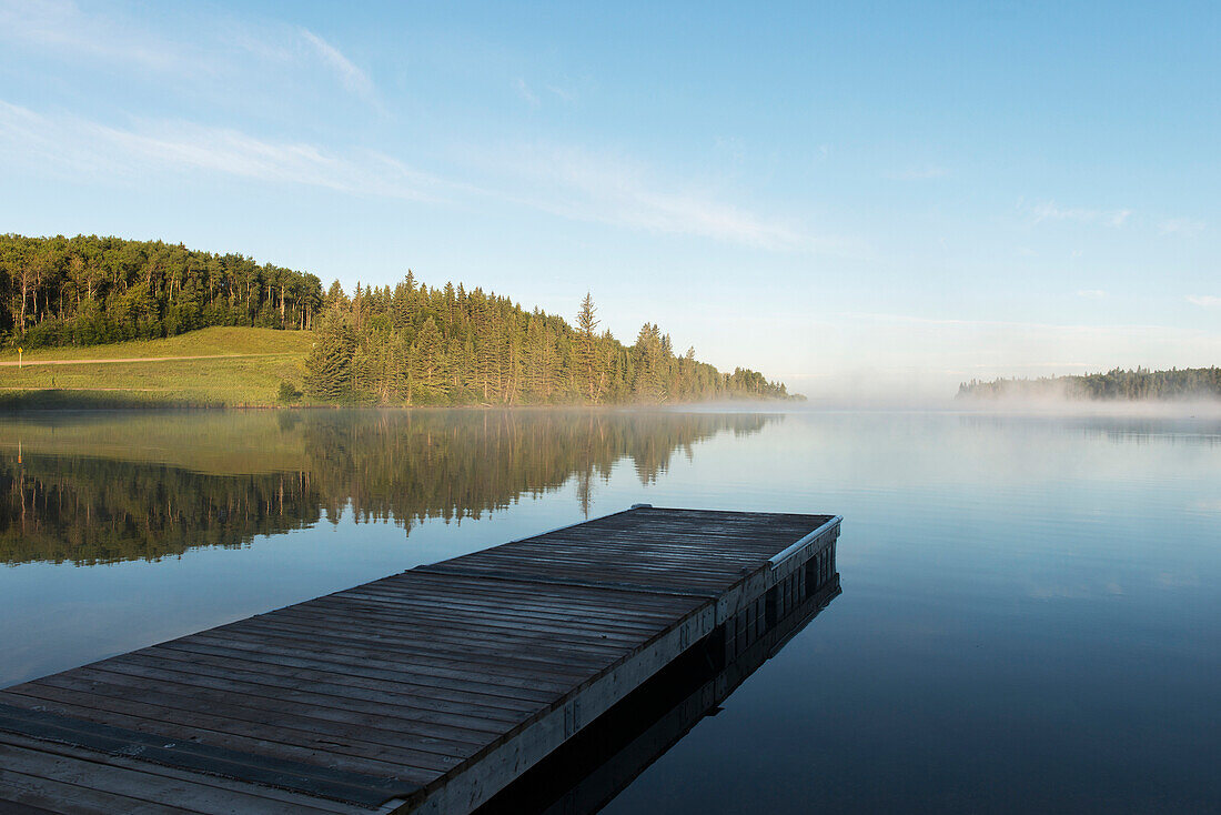 'A wooden dock leading out to a misty tranquil lake in Riding Mountain National Park; Wasagaming, Manitoba, Canada'