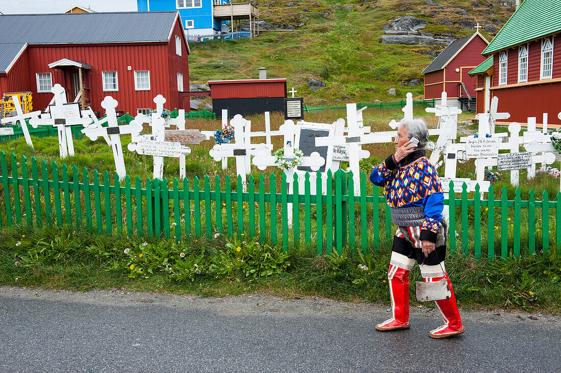 Frau in traditioneller Kleidung telefoniert mit einem Handy vor einem Friedhof mit Holzkreuzen, Nuuk, Grönland