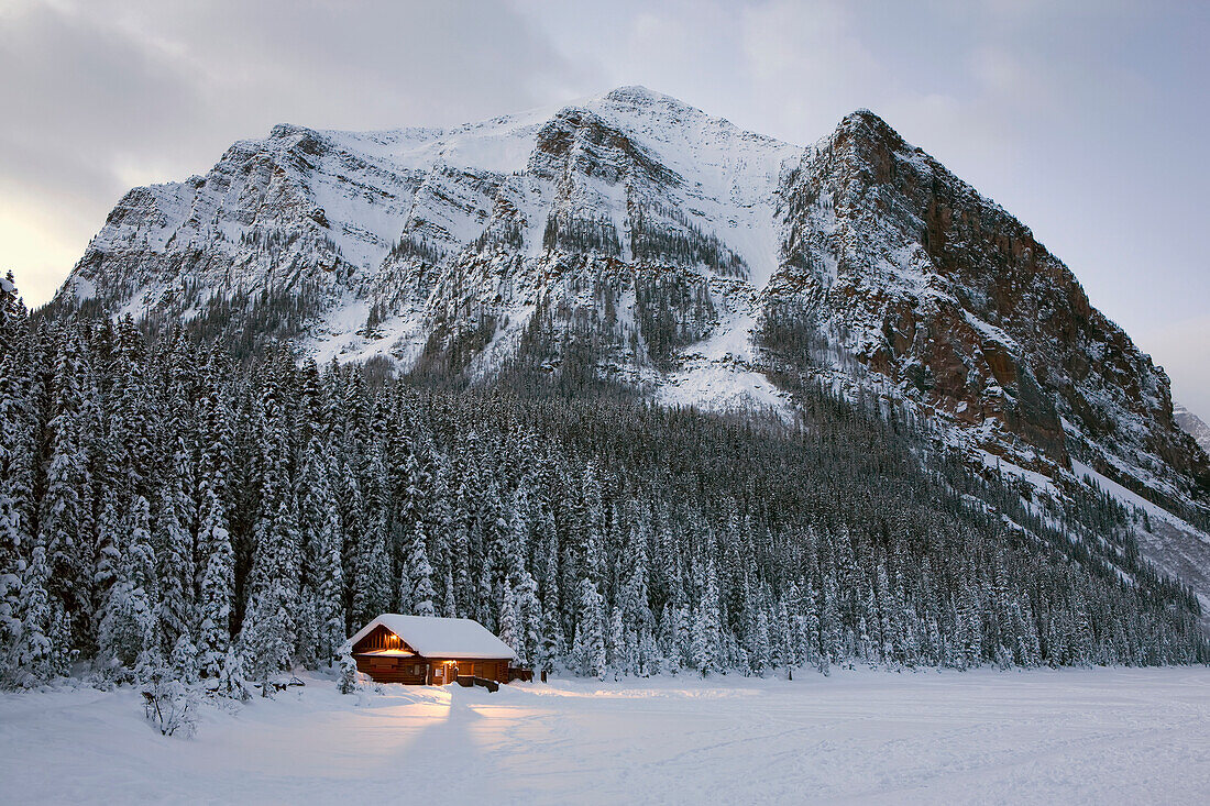 'A snow covered log cabin on a lakeshore with lights at dusk surrounded by snow covered evergreen trees and mountains;Lake louise alberta canada'