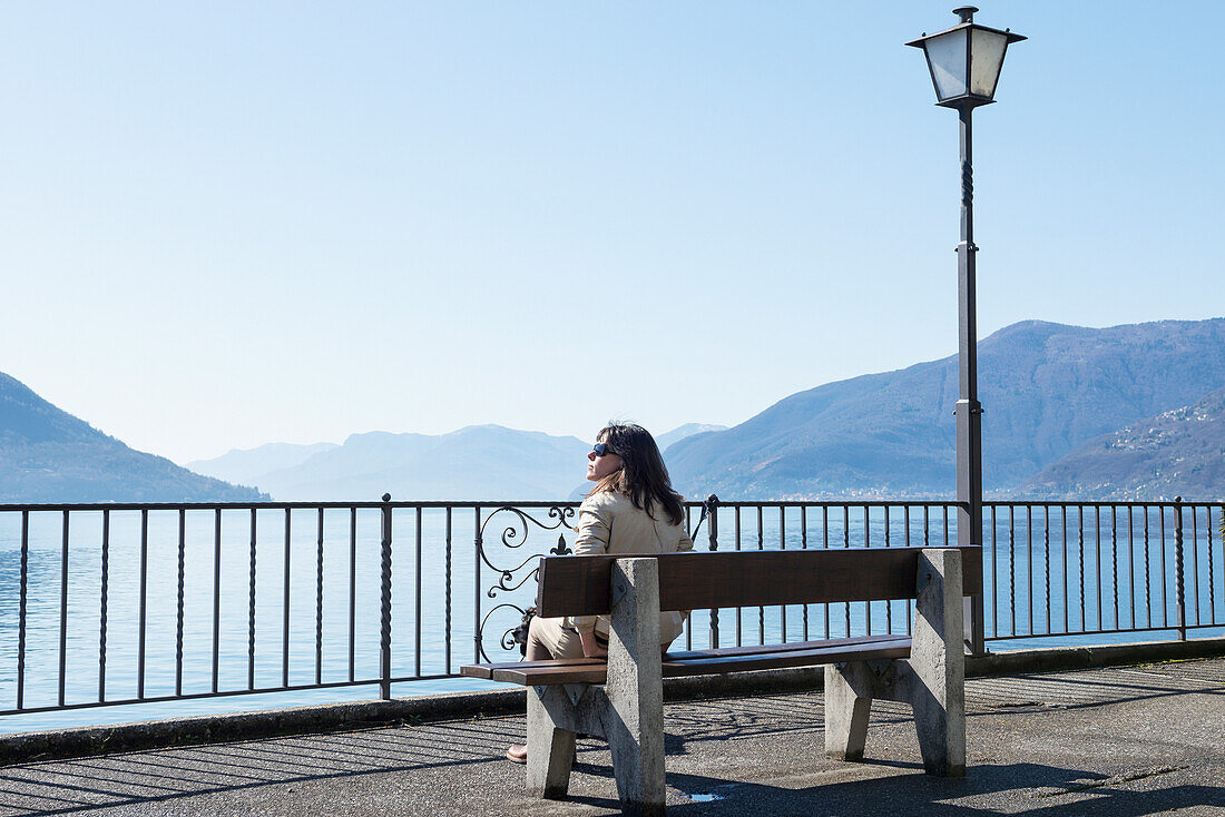 'A Woman Sitting On A Bench Overlooking Lake Maggiore; Brissago, Ticino, Switzerland'