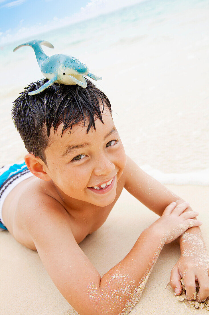 'A Boy Laying On The Sand At The Water's Edge With A Toy Whale On His Head; Kailua, Oahu, Hawaii, United States Of America'