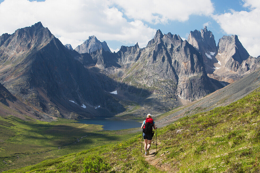 'Female hiker walking on mountain trail with mountain lake, and valley with clouds and blue sky; Yukon, Canada'