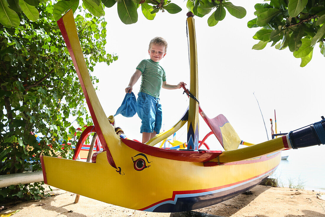Young boy climbing on boat, traditional fisherboat, 3 years old, beach, eye, colorful boat, prahu, play, family travel, Indian Ocean, parental leave, MR, Sanur, Bali, Indonesia