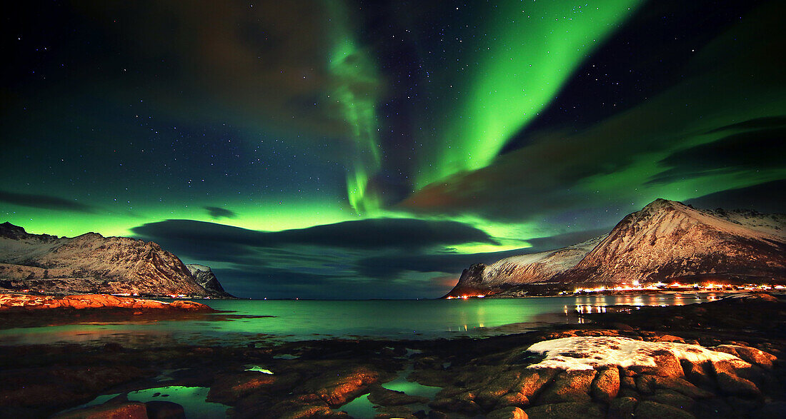Aurora borealis reflected in the sea, with clouds seen from a fjord, green sky with big mountain in background