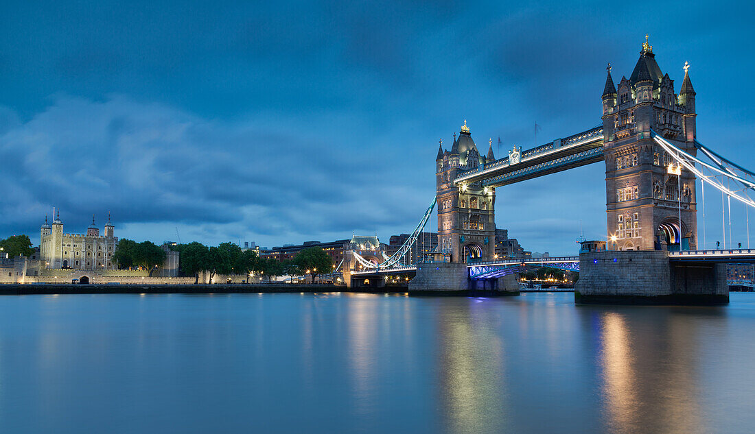 The Tower Bridge and the Tower of London are reflected in the calm waters of the Thames in the early evening lights.