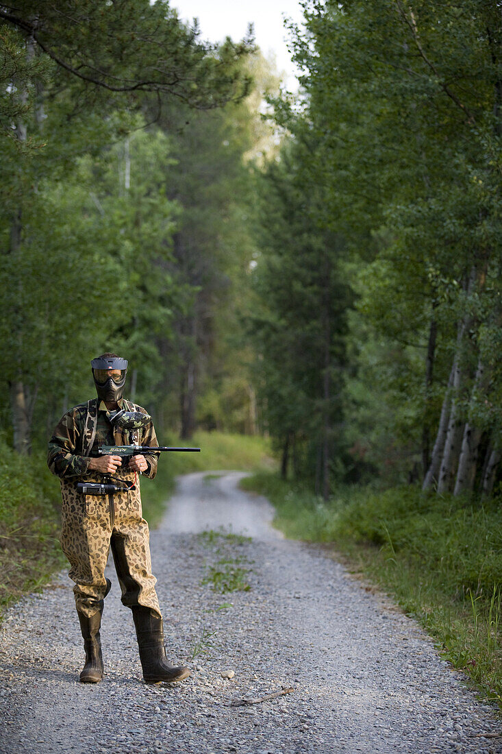 A man dressed in paintball garb poses on a forest trail.
