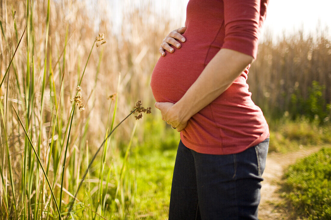 A pregnant woman holds her belly for a portrait in front of tall beach grasses on a sunny day in California.