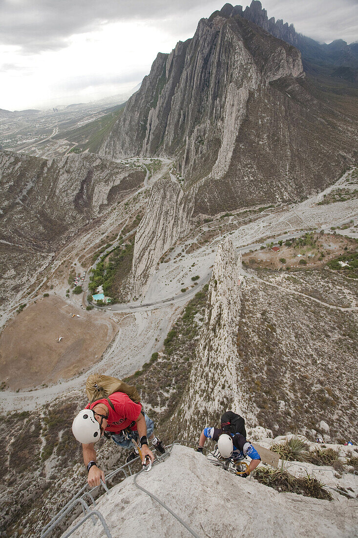 Two men with backpacks climbing a via ferrata in La Huasteca with some mountains in the background. La Huasteca, Nuevo Leon, Mexico.