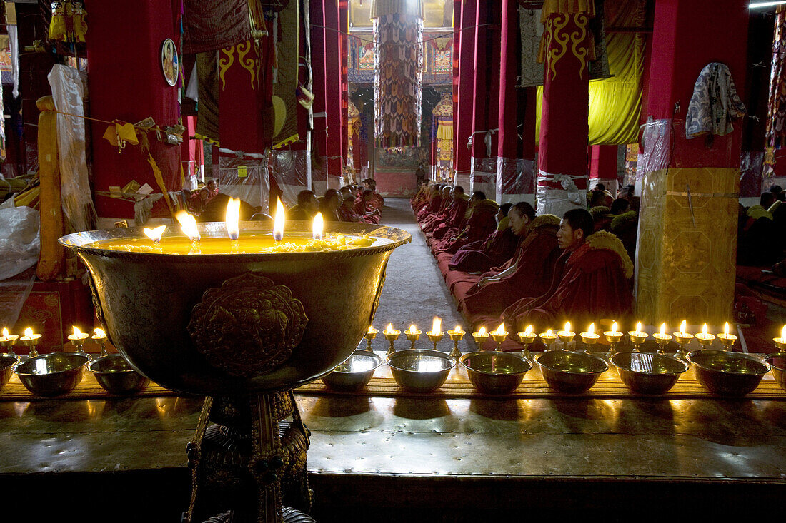 The main prayer hall of Drepung monastery embellished with red columns, victory banner, thankas and monastic paintings on wall.  Seen here monks praying.  Tibet.