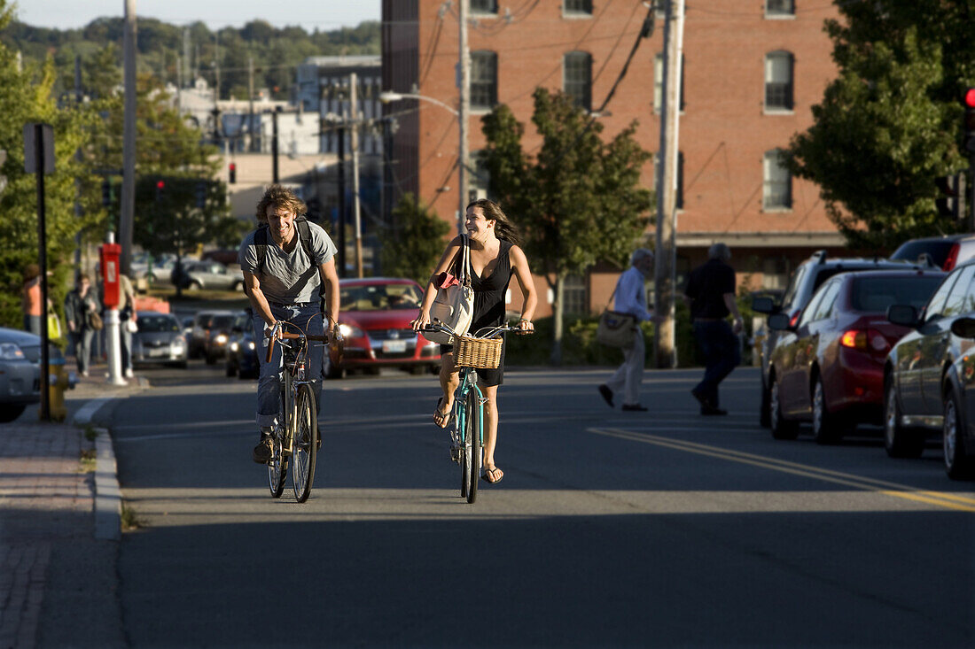 A young man and woman smile as they ride their bikes up a busy street on a sunny afternoon in Portland, ME.
