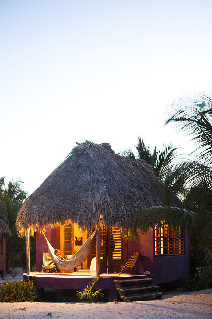 Ambergris Cay, Belize. A woman sits in a hammock on the porch of her thatch cabana at a luxury hotel in Belize.
