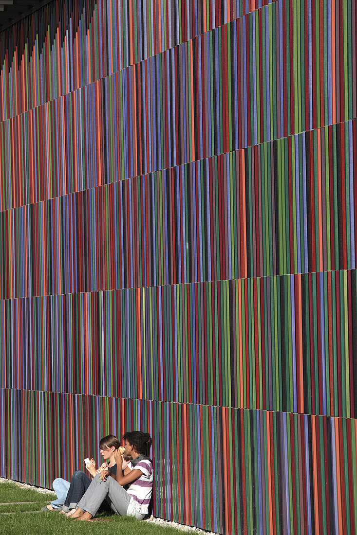 Two girls sit against a colorful wall outside the Brandhorst Museum in Munich, Bavaria, Germany.
