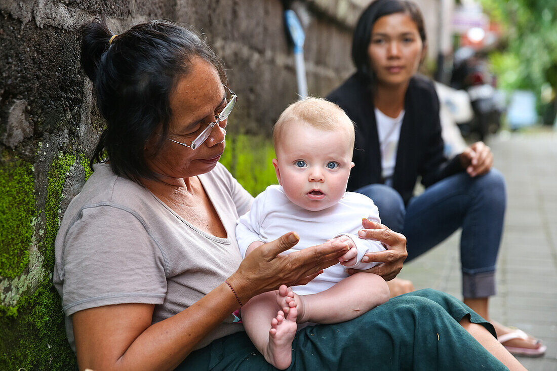 Balinesische Frau spielt mit westlichem Baby, Mädchen 5 Monate alt, sitzt auf Schoß, interkultureller Kontakt, Elternzeit in Asien, Europäer, Deutsche, Westler, Familie, MR, Ubud, Bali, Indonesien