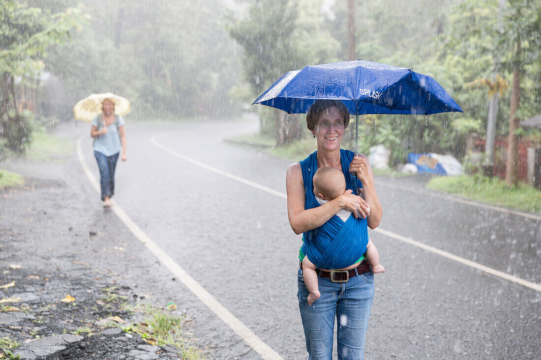 Tropical rain, monsun, heavy rainfall, tourists walking along a road, German mother with her baby, wrap, baby sling, 5 months old, family travel in Asia, parental leave, German, European, MR, Munduk, Bali, Indonesia