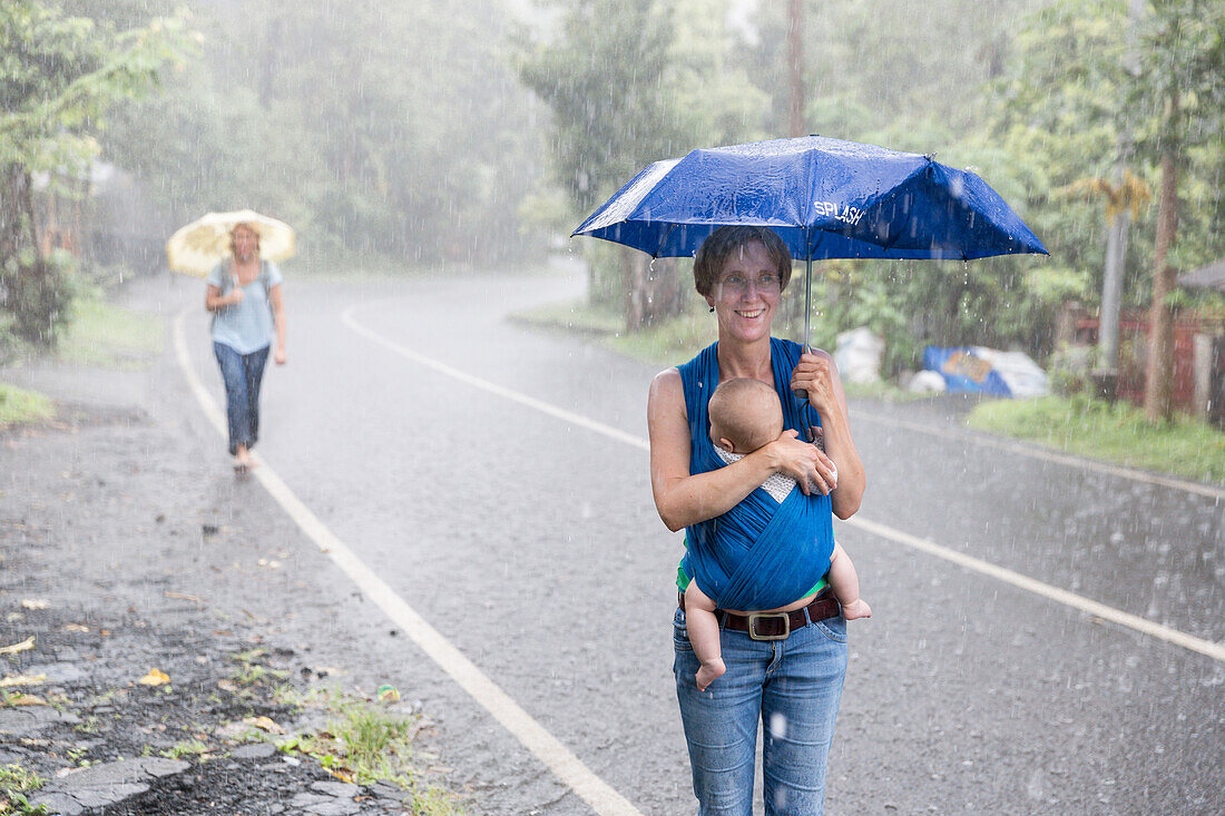 Tropischer Regen, Sturzregen, Platzregen, Monsunregen, Monsun, Urlauber, Touristen, Straße, deutsche Mutter mit Baby im Tragetuch, Regenschirm, lachen, Mädchen 5 Monate alt, Elternzeit in Asien, Europäer, Deutsche, Westler, Familie, MR, Munduk, Bali, Indo