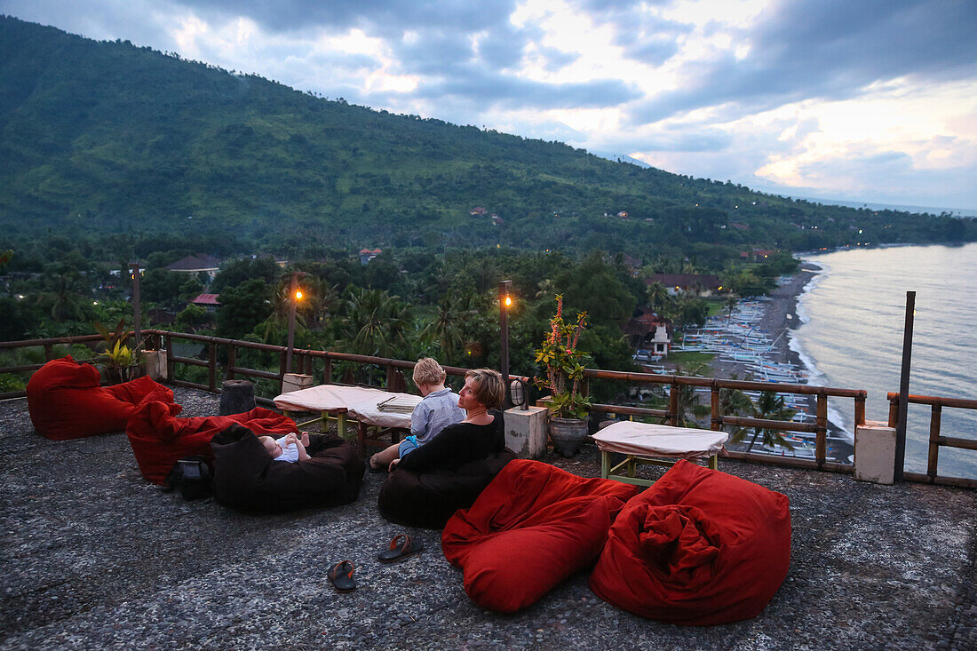 German family, dinner, mother with her two children, restaurant high above the beach, terrace, sea, shore, vulcano, red cushions, dusk, evening, boy 3 years, daughter 5 months, baby, family travel in Asia, parental leave, German, European, MR, Amed, Bali,