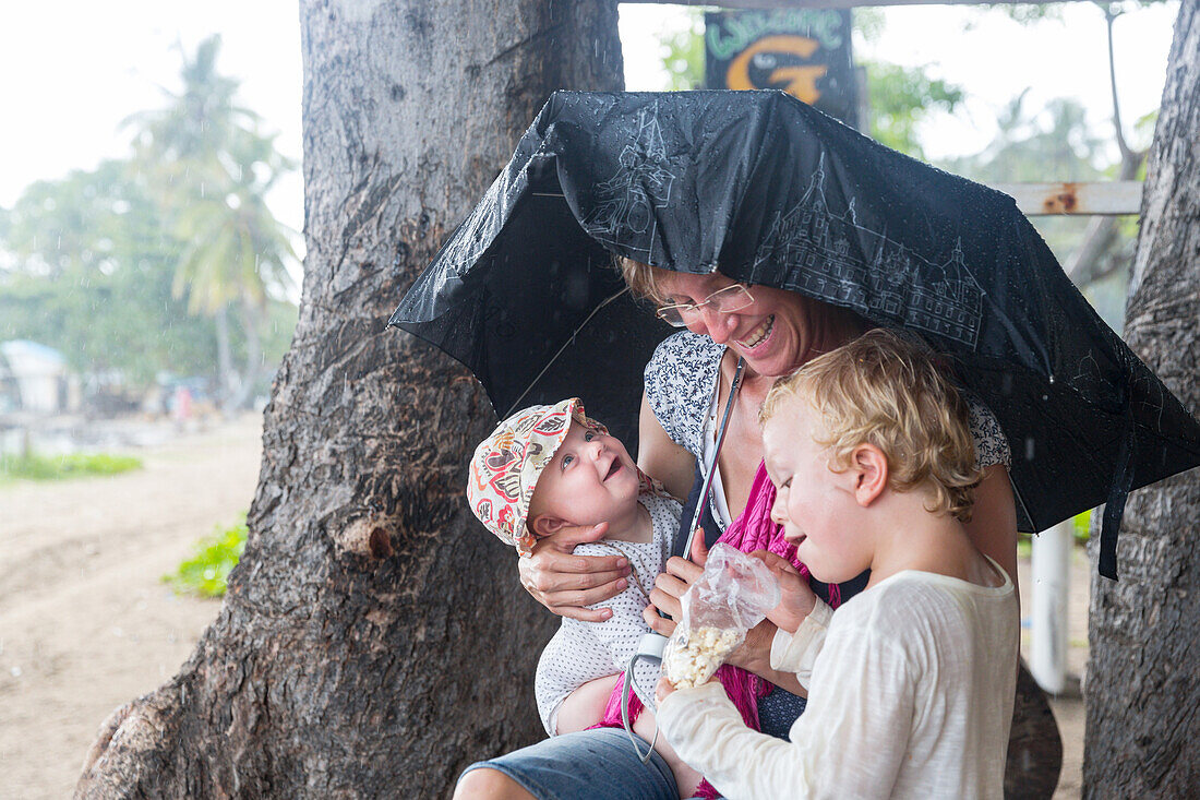 Family in the rain, mother with her two children under umbrella, baby girl 5 months, boy 3 years old, parental leave, German, Gili Isles, Lombok, Indonesia